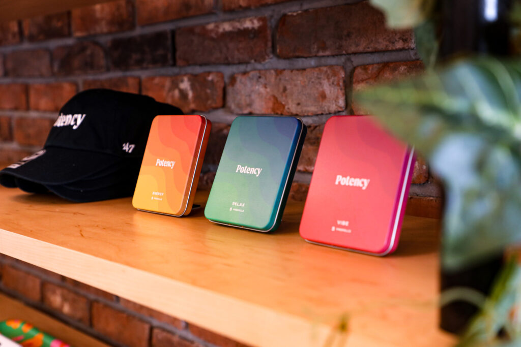 A wooden shelf displaying three colorful rectangular tins labeled "Potency" with different labels—"Energy," "Relax," and "Vibe"—alongside black caps embroidered with the word "Potency." The background features a rustic red brick wall, and part of a green plant is visible on the right side.